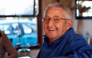 A smiling senior man, happy as result of successful long-distance caregiving, is sitting at a table and looking over his shoulder.
