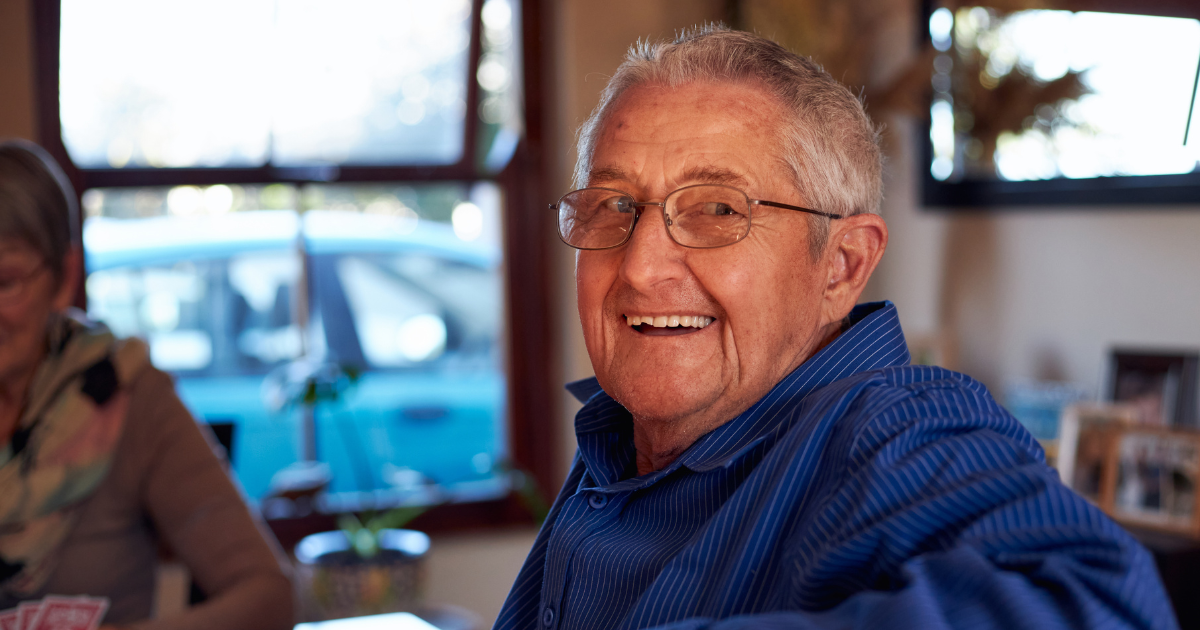 A smiling senior man, happy as result of successful long-distance caregiving, is sitting at a table and looking over his shoulder.