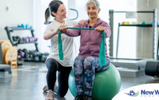 A care provider helps an older woman exercise with a resistance band and an exercise ball, representing how exercise can help with senior fall prevention.