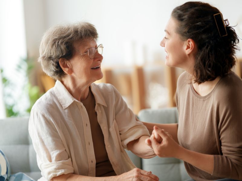 A smiling female caregiver in a brown shirt holds the hand of an elderly woman with glasses, who is smiling back warmly. They are seated in a living room, illustrating Compassionate Alzheimer's and Dementia Care in Palos Verdes, CA