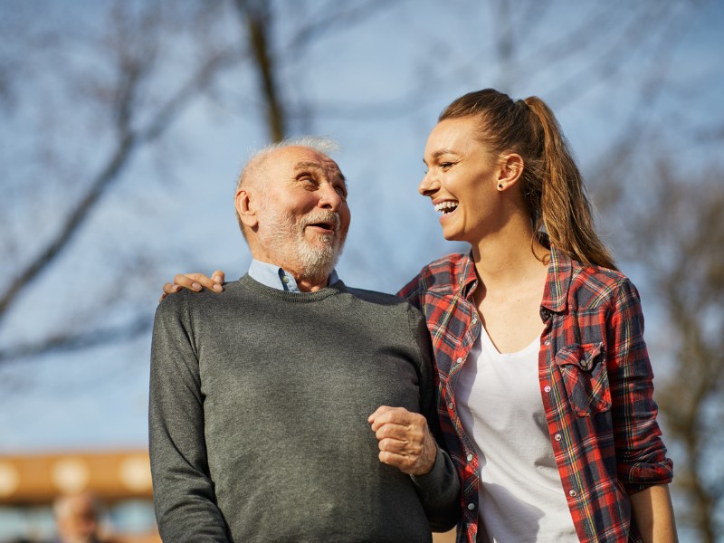 A joyful outdoor scene of a younger female caregiver laughing with an elderly man during a walk. Their genuine connection and the man's happy expression highlight the positive impact of specialized Alzheimer's and Dementia Care in Santa Monica, CA