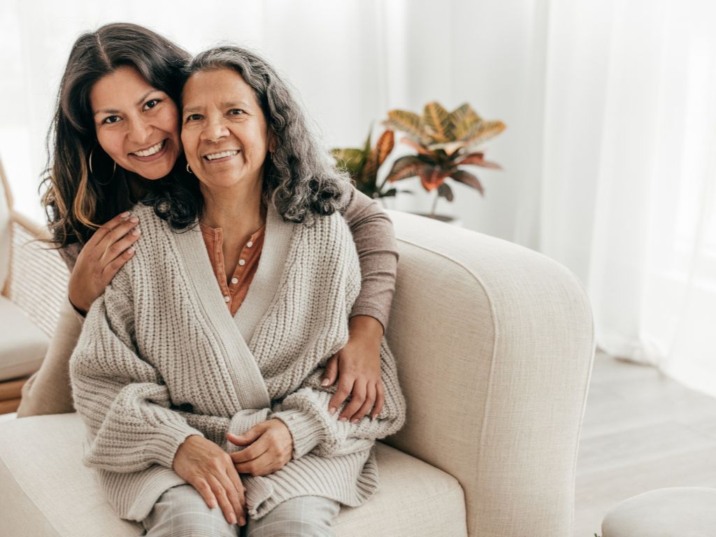 A smiling senior woman sitting on a couch with her caregiver embracing her, representing compassionate Alzheimer’s and dementia care in Torrance.