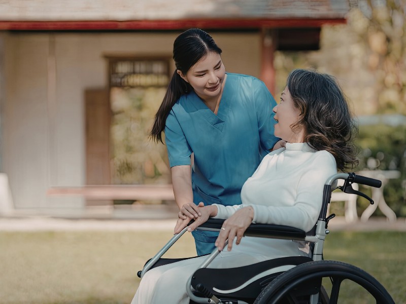 A compassionate female caregiver in blue scrubs gently holds the hand of an elderly woman seated in a wheelchair outdoors. Their peaceful and supportive interaction illustrates the empathy of our Hospice Support in Hermosa Beach, CA services.