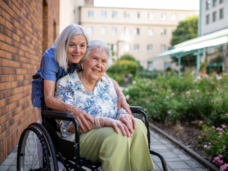 A female caregiver with silver hair affectionately hugs a smiling elderly woman in a wheelchair outdoors on a paved path. The warm embrace and garden setting illustrate the compassionate nature of our Transitional Care in Hermosa Beach, CA programs.