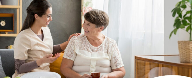 Smiling caregiver with her hand on an older woman’s shoulder as they share tea on the couch, showing home care vs assisted living