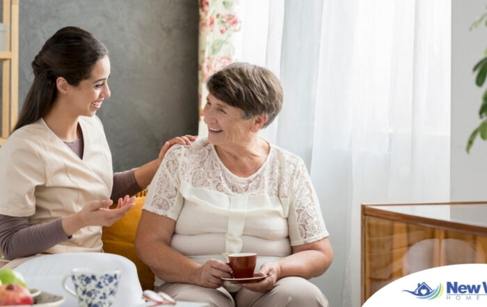 Smiling caregiver with her hand on an older woman’s shoulder as they share tea on the couch, showing home care vs assisted living